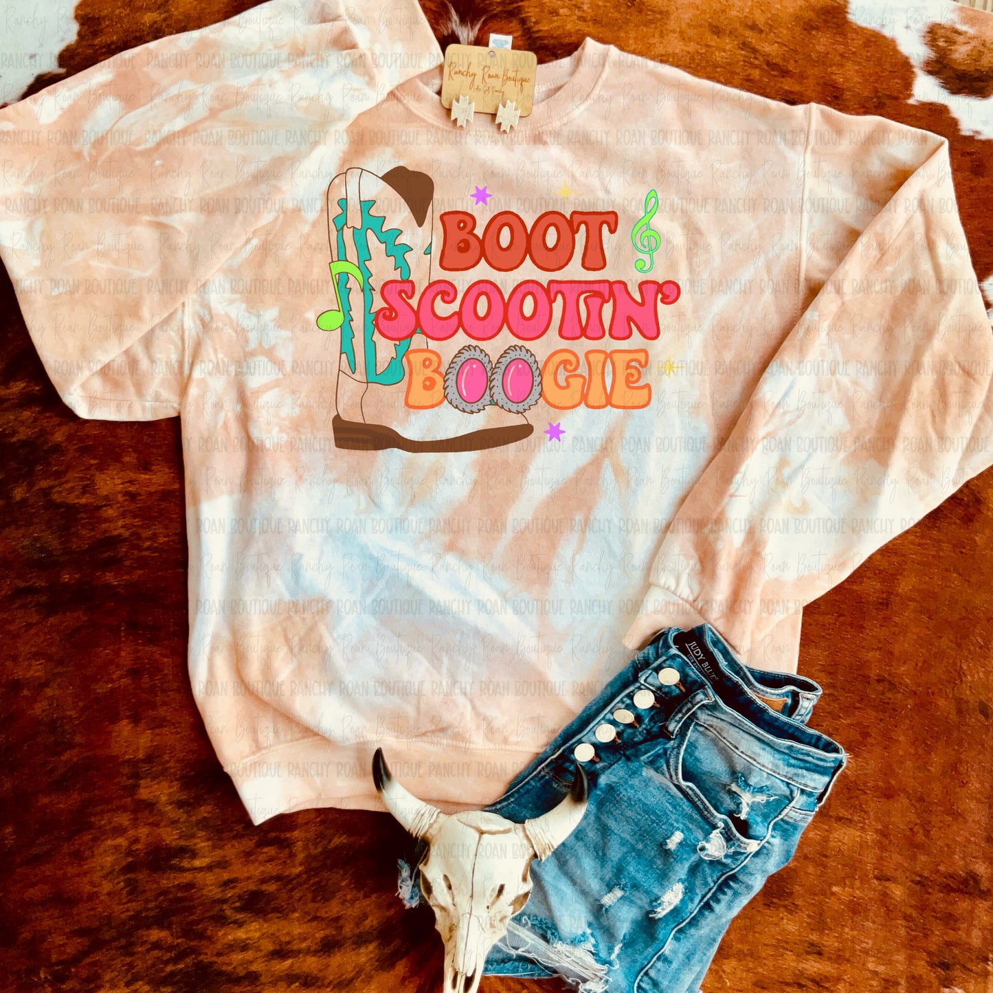 Hand-dyed white sweatshirt in tan and brown tones featuring colorful Boot Scootin’ Boogie Western graphic, styled with distressed denim shorts and cow skull decor.