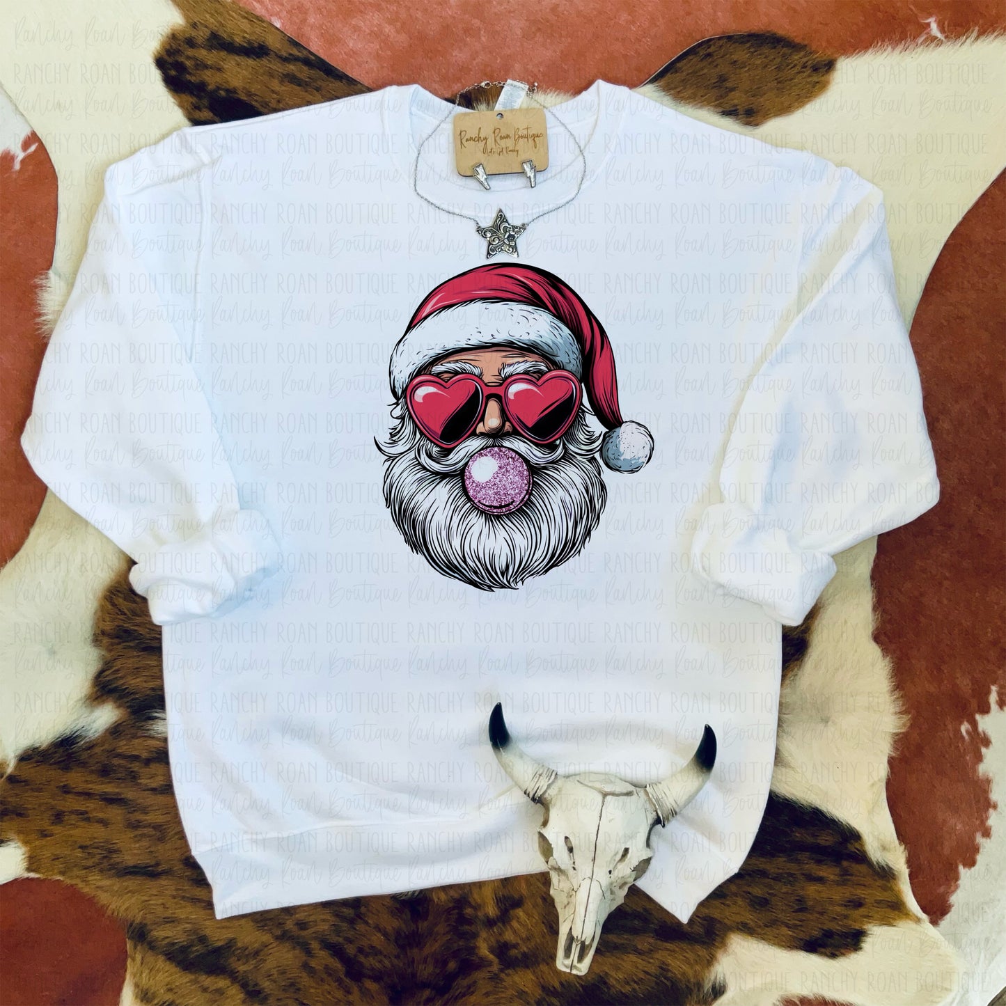 White sweatshirt featuring a playful graphic of Santa with heart-shaped sunglasses and blowing a bubblegum bubble, styled with Western-themed decor.