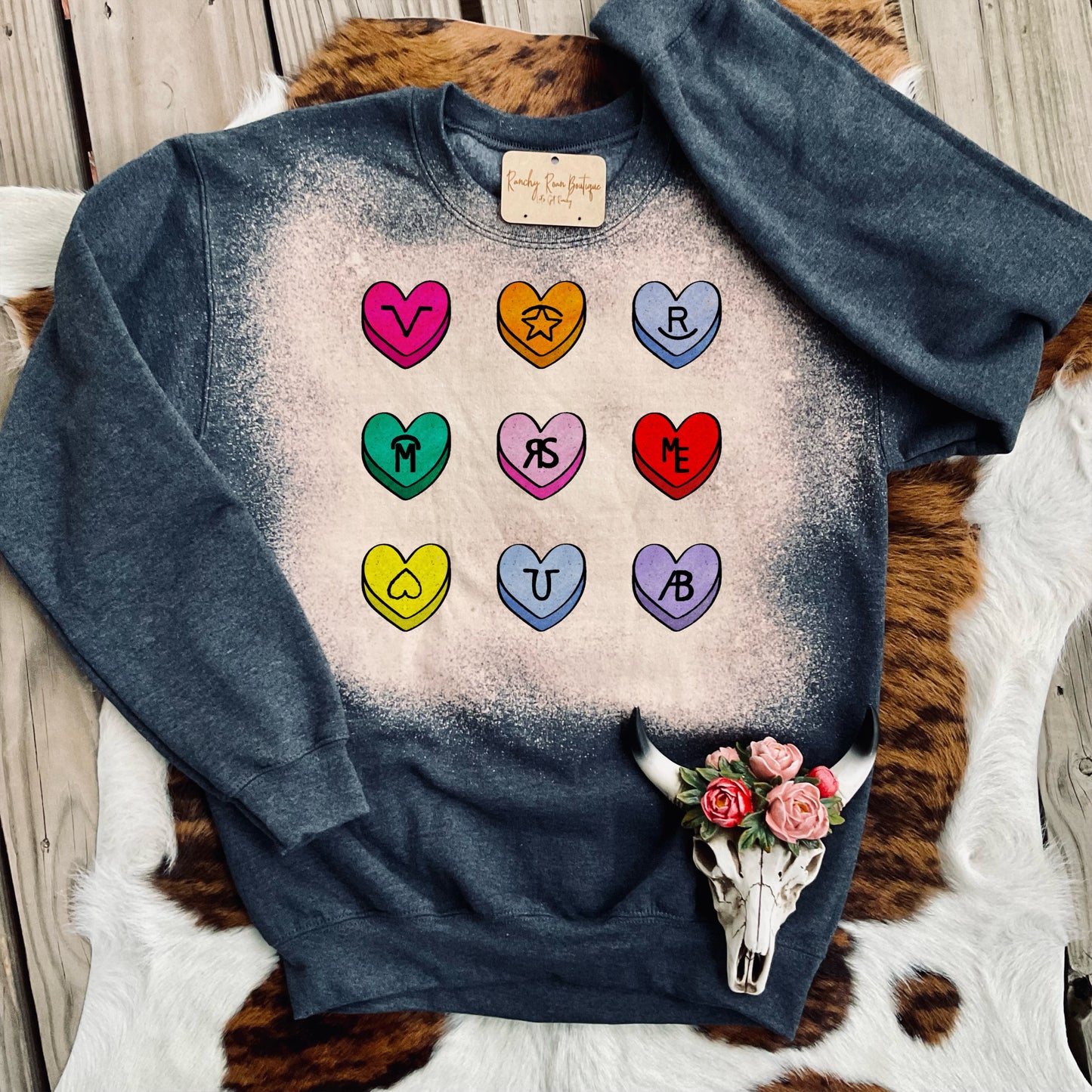 A fun sweatshirt featuring colorful candy-heart designs with Western icons, displayed with a cowhide backdrop.