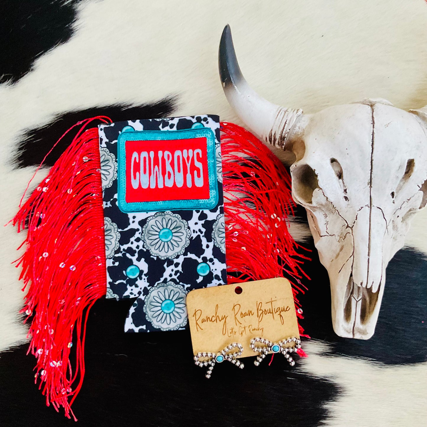 Cow print koozie with “Cowboys” text patch and red fringe, accented by turquoise concho detailing.
