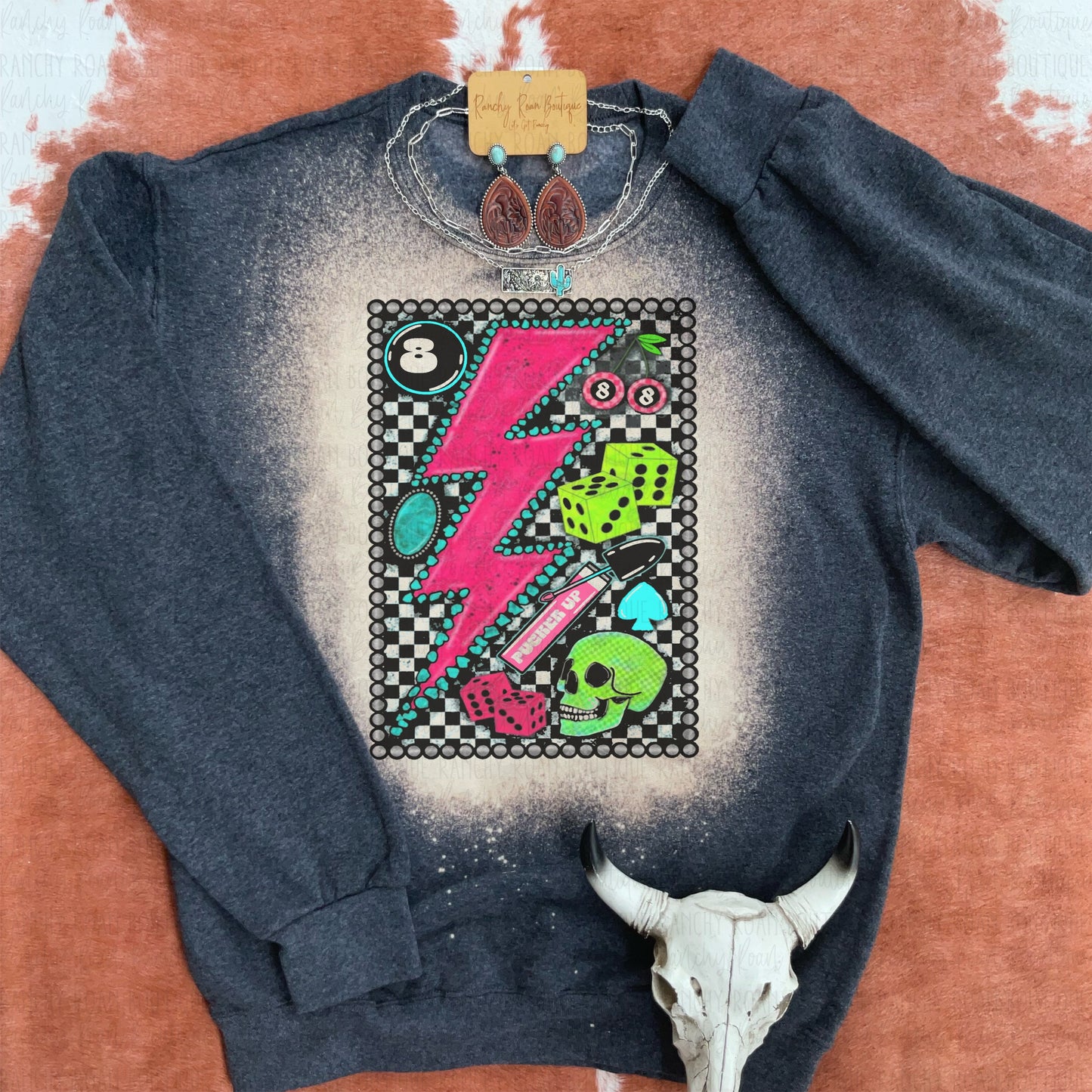 Dark graphic sweatshirt with neon lightning bolt, skull, dice, and checkerboard background, displayed on a cowhide with a decorative cow skull.