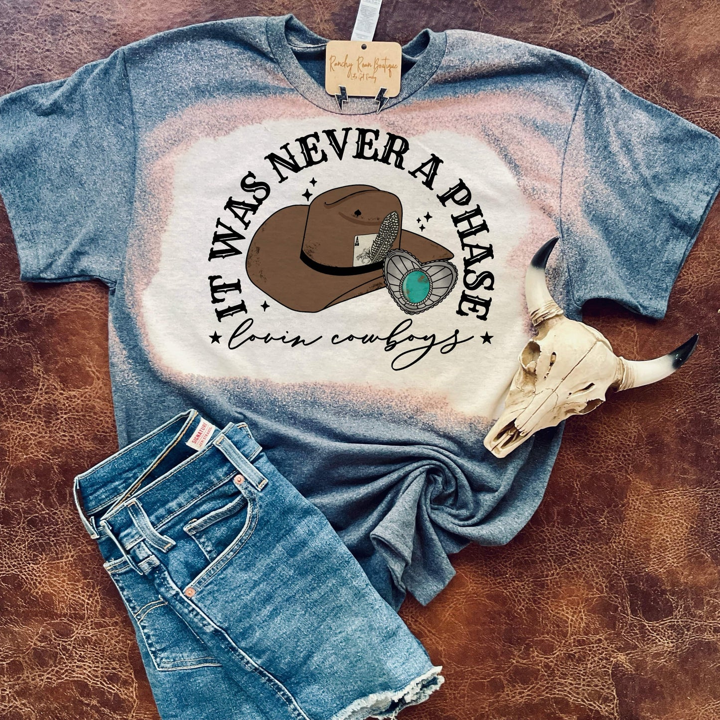 It_Was_Never_a_Phase_Lovin_Cowboys_Bleached_Tee: A bleached tee from Ranchy Roan Boutique featuring the text “It Was Never a Phase Lovin’ Cowboys” with a cowboy hat and turquoise ring graphic. The shirt has a unique bleached pattern with shades of blue and cream, styled with denim shorts and a cow skull accessory.