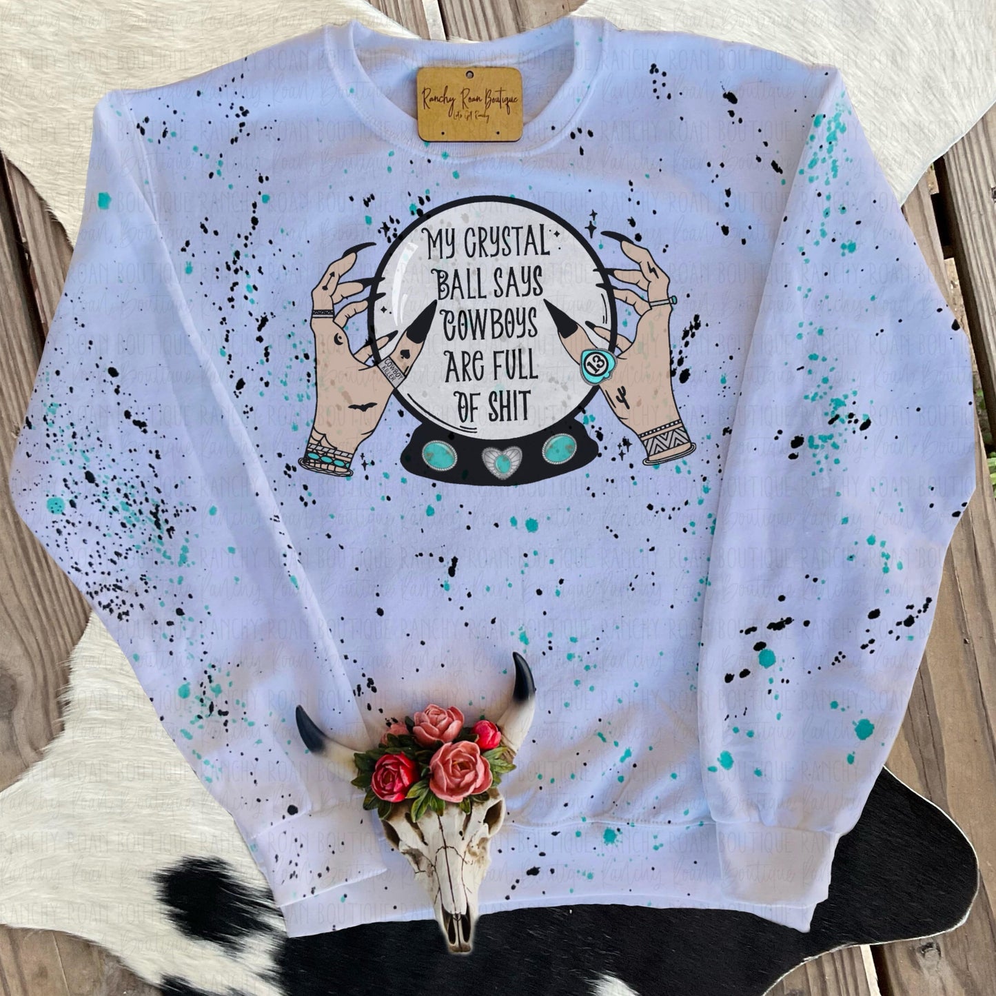 White sweatshirt with turquoise and black paint splatter, featuring “My Crystal Ball Says Cowboys Are Full of Shit” Western Halloween-themed sublimation graphic, displayed flat.