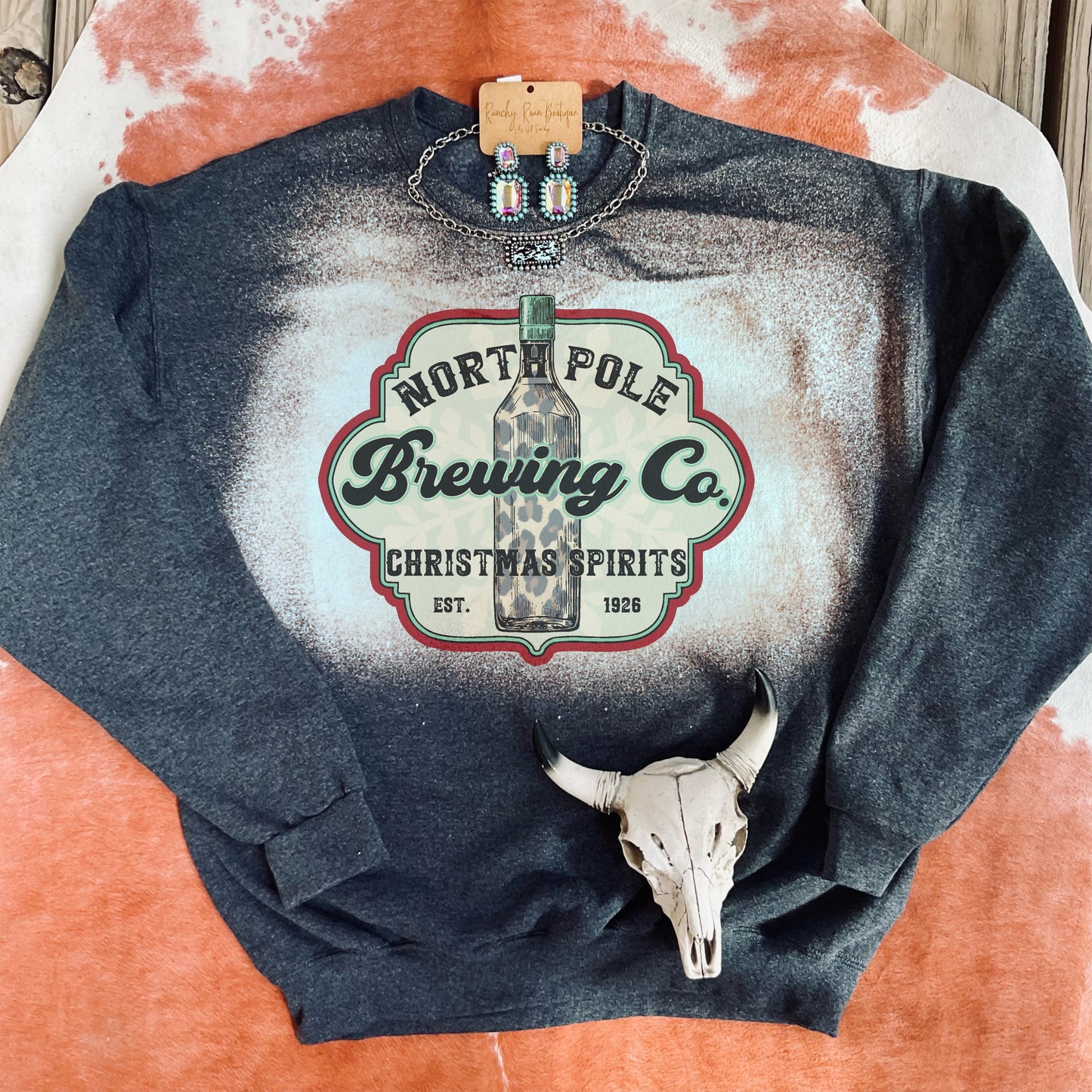 A black Heather unisex sweatshirt with a bleach splatter design and a sublimated “North Pole Brewing Co. Christmas Spirits” graphic, featuring a leopard print bottle.