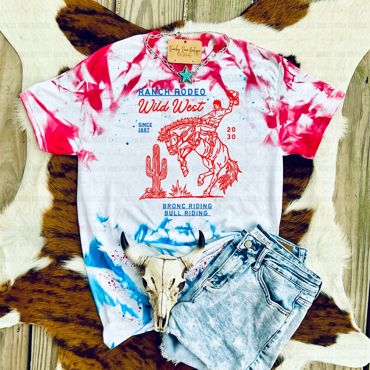 Patriotic Western bronc riding graphic tee with red and blue tie-dye, styled on a cowhide background.
