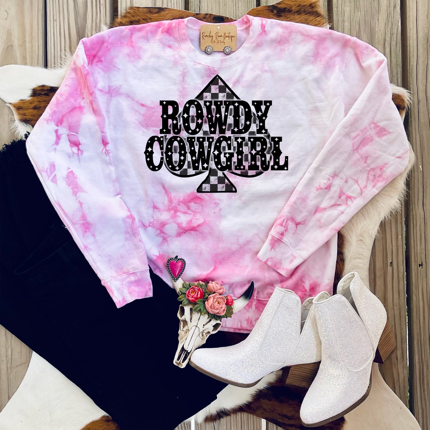 A vibrant pink tie-dye long sleeve with bold “Rowdy Cowgirl” spade graphic, styled with white boots and accessories.