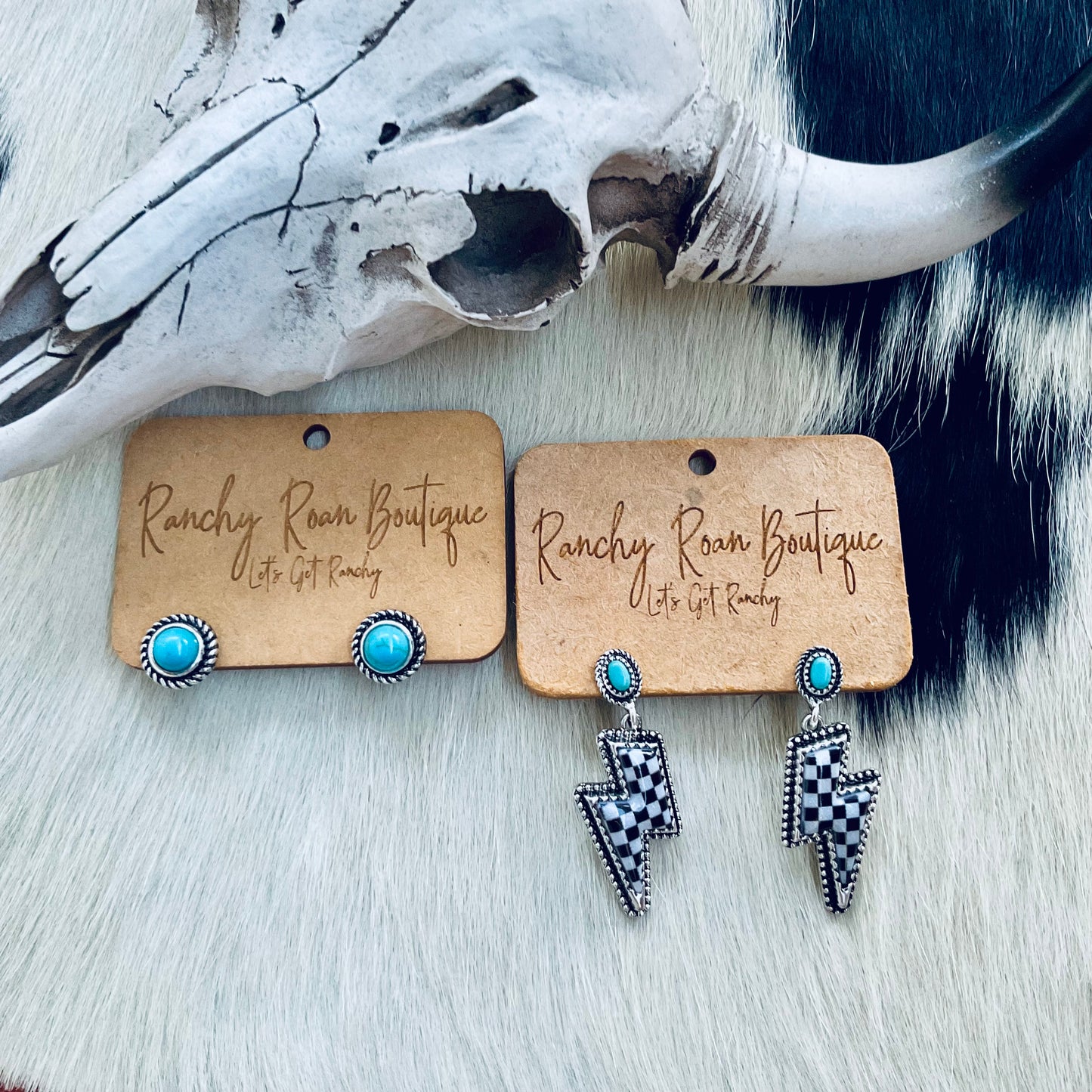 A pair of turquoise stud earrings and black-and-white checkered lightning bolt earrings, both displayed on Ranchy Roan Boutique cards, set against a cowhide background with a decorative cow skull.