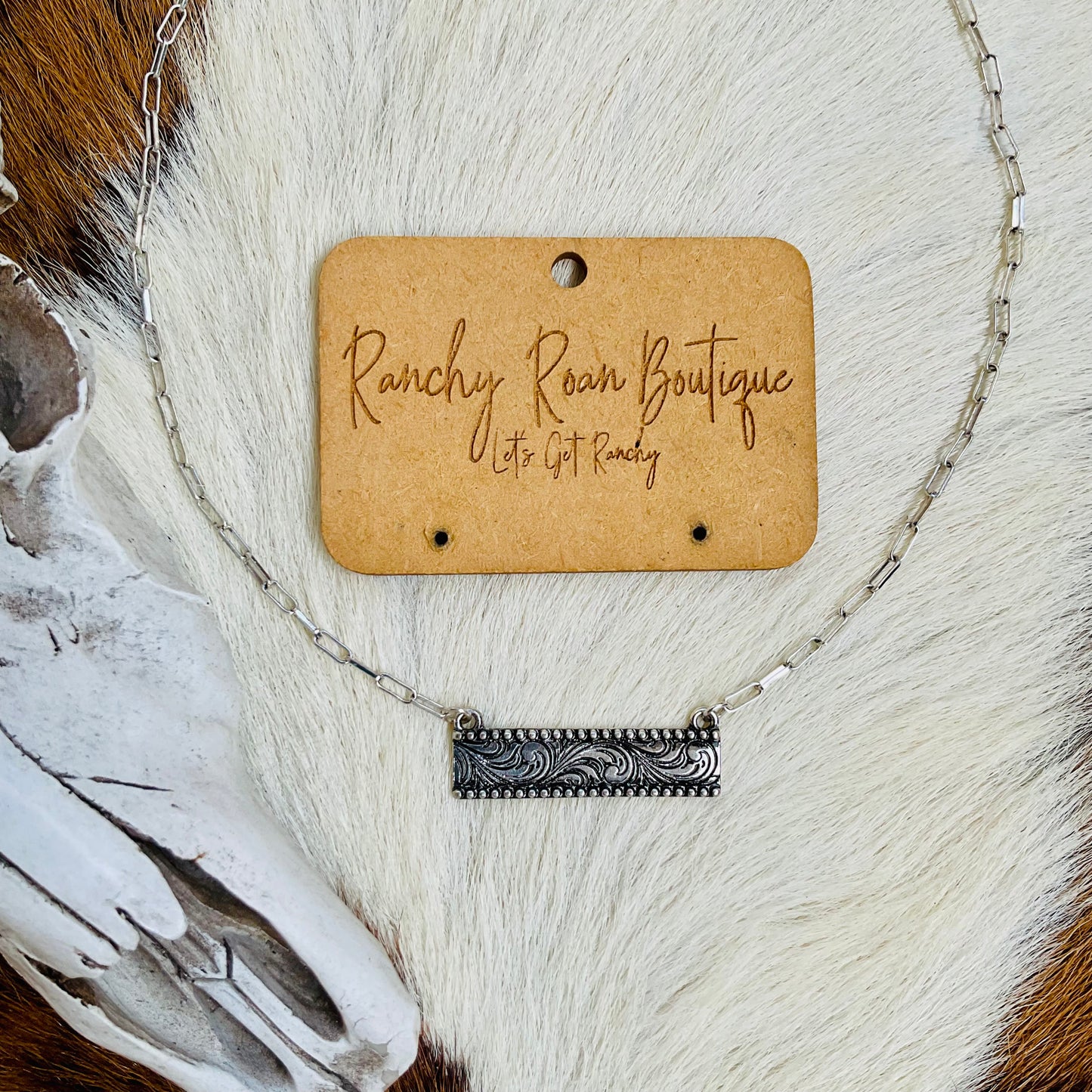 Dainty necklace laid flat with a Western skull prop, highlighting the engraved details on the burnished silver bar pendant.