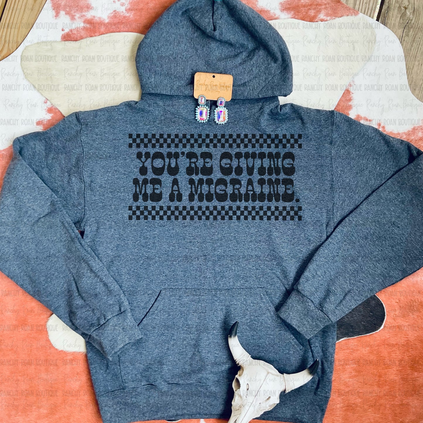 Dark Heather hoodie with bold black checkered graphic that reads ‘You’re Giving Me a Migraine’ – styled with beaded earrings and cow skull on cowhide background”