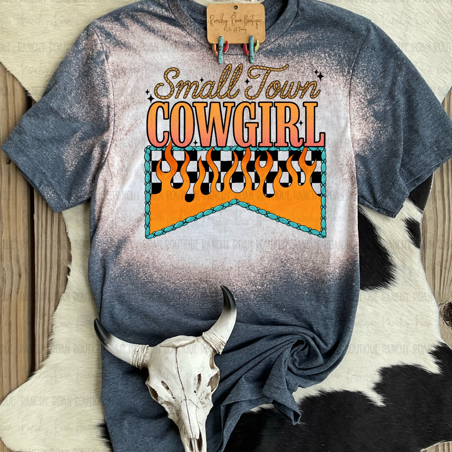 Western bleach graphic tee with “Small Town Cowgirl” sublimated design featuring cow print flames and turquoise rope, styled with skull and earrings on cowhide