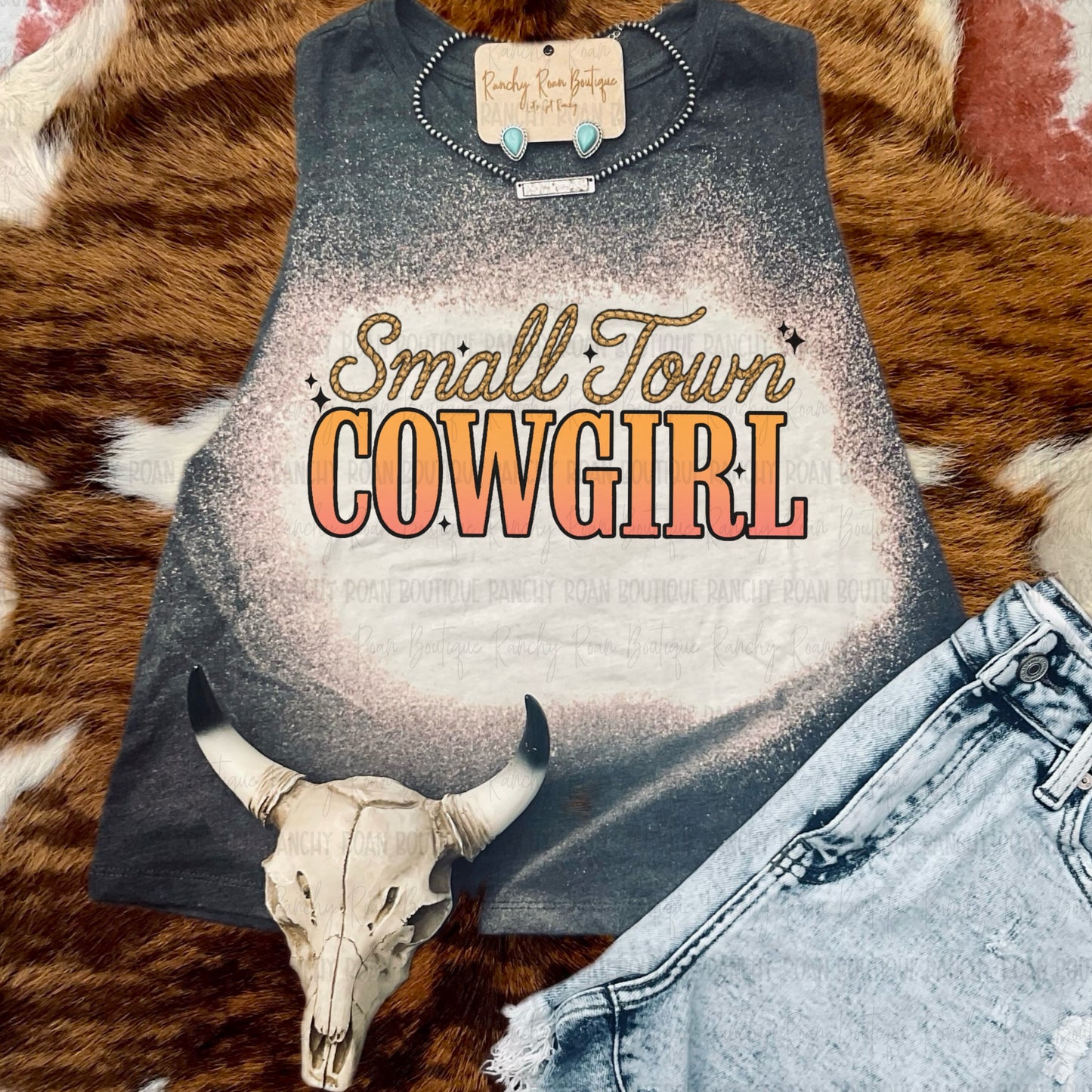 Western bleach muscle tank with sublimated “Small Town Cowgirl” text in peach and gold, styled with turquoise jewelry and distressed shorts on cowhide