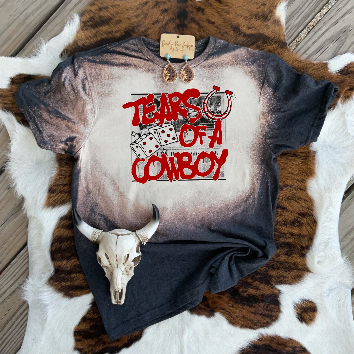 Bleach-washed unisex Western tee with sublimated “Tears of a Cowboy” graphic featuring dice, newspaper print, and a horseshoe, styled on cowhide