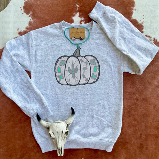 A light heather gray sweatshirt from Ranchy Roan Boutique featuring a large pumpkin graphic with Aztec patterns and turquoise accents, enhancing its Southwestern flair. Styled with a cow skull accessory to complement the Western theme.