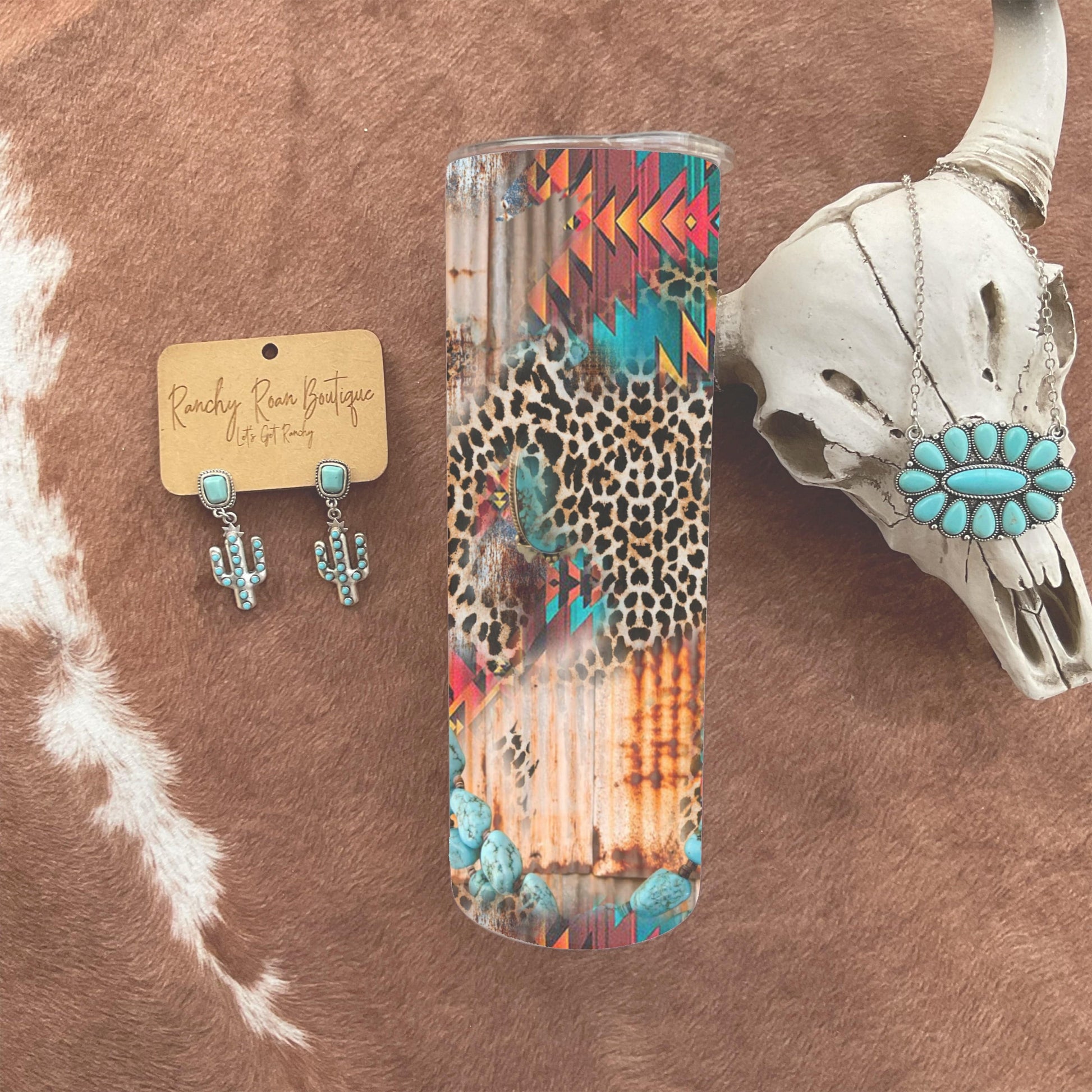 Aztec Turquoise Leopard 20oz Skinny Tumbler featuring bold Aztec patterns, turquoise accents, and a leopard print on a rusted metal background, styled with Western accessories.
