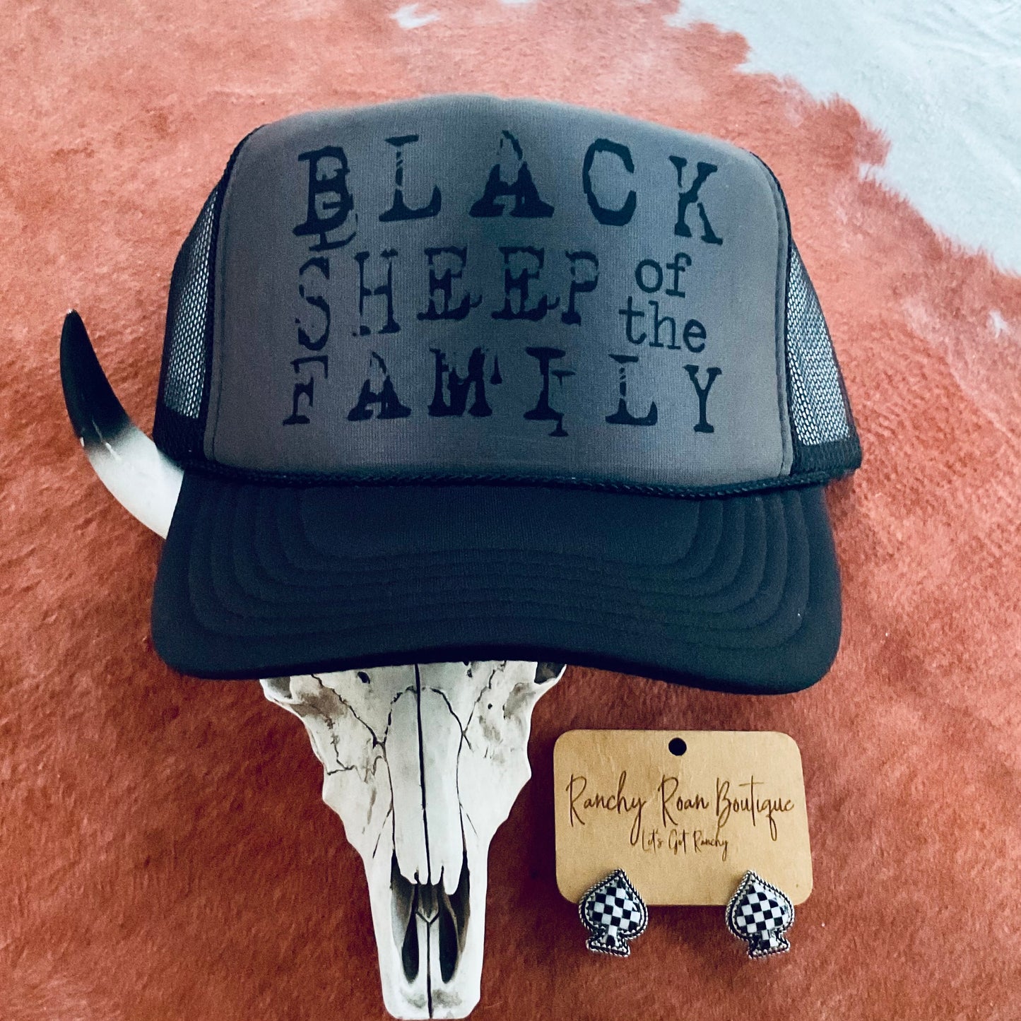 Full display of the “Black Sheep of the Family” trucker hat resting on a cowhide with decorative earrings, featuring the structured foam front and mesh back.