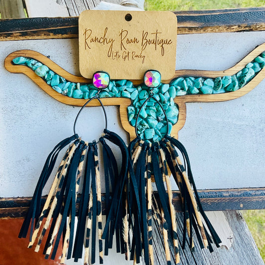  Black teardrop earrings with iridescent glass stone studs and black/leopard leather tassels, displayed on a Ranchy Roan Boutique card against a wooden turquoise horn background.