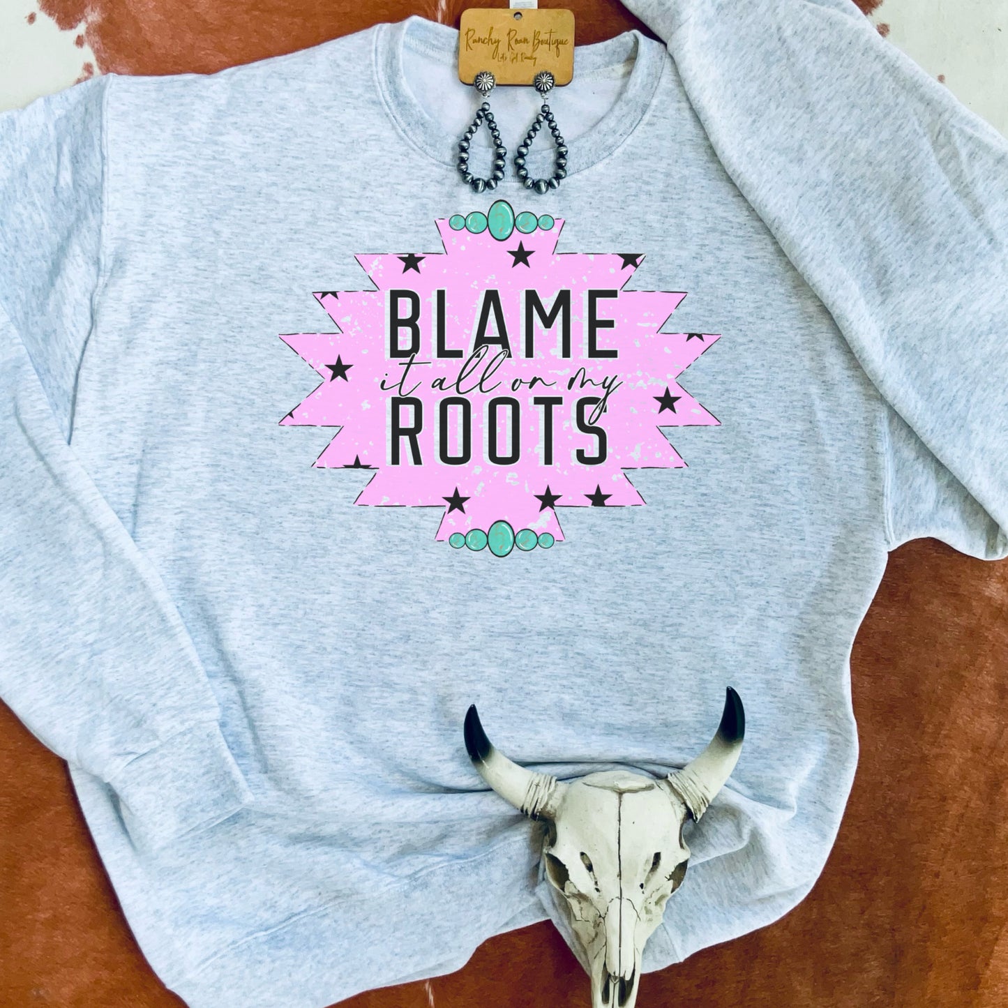 Gray sweatshirt featuring the pink “Blame It All On My Roots” graphic with black stars and turquoise accents, displayed with Western decor.