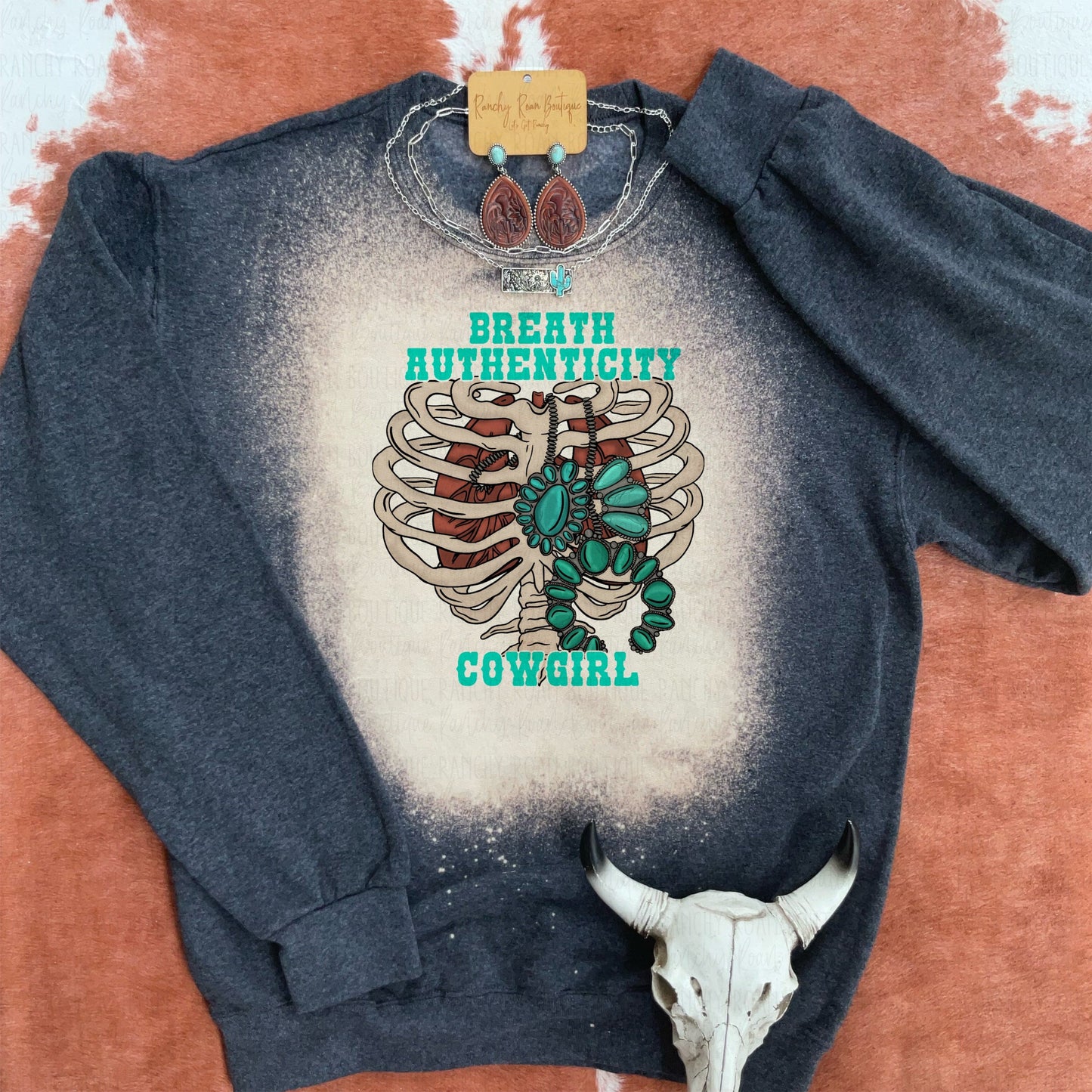 “Breathe Authenticity Cowgirl” Graphic Sweatshirt with a ribcage and turquoise jewelry design, displayed with western accessories on a rustic background.