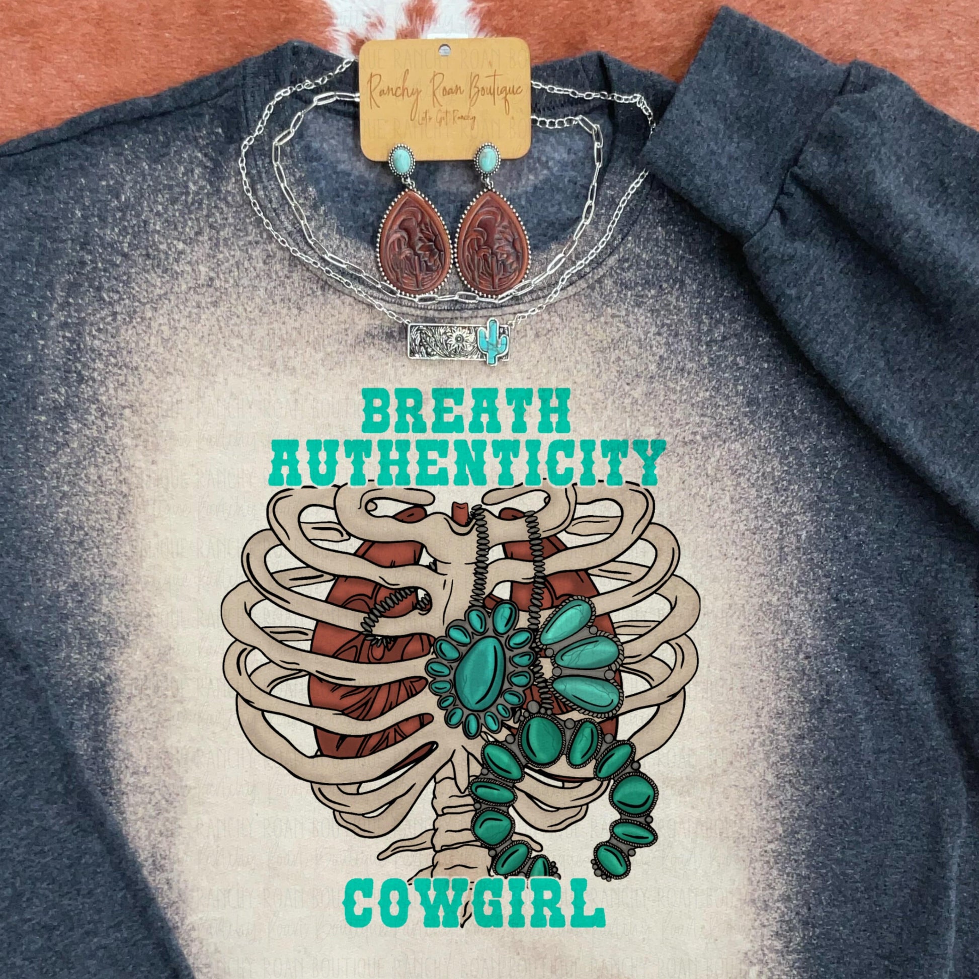 “Breathe Authenticity Cowgirl” Graphic Sweatshirt with a ribcage and turquoise jewelry design, displayed with western accessories on a rustic background.