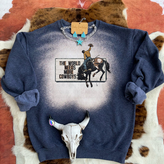 Dark unisex crew sweatshirt with a graphic of a cowboy riding a bucking bronco, featuring the text “The World Needs More Cowboys” and a bleach splatter design.
