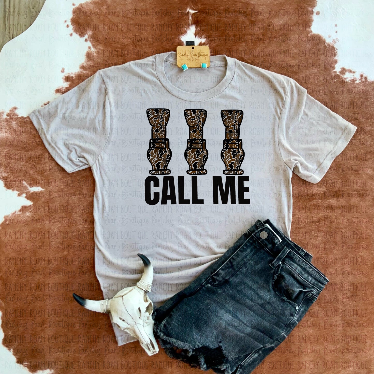 Call Me Camo Hunting Western Graphic Tee displayed with a decorative skull and turquoise earrings on a cowhide rug.