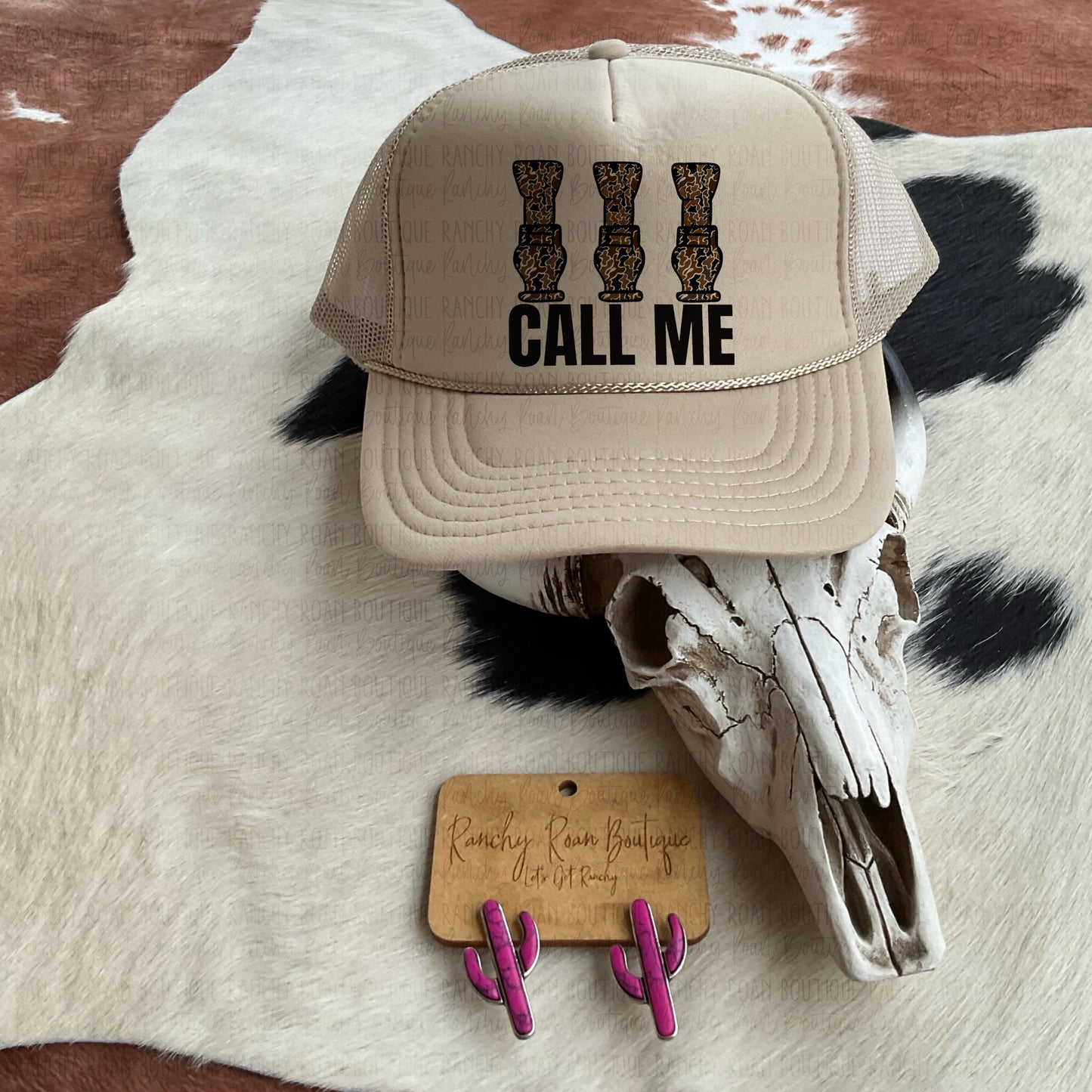 Call Me Camo Hunting Western Trucker Hat displayed with a decorative skull and pink cactus earrings on a cowhide rug.