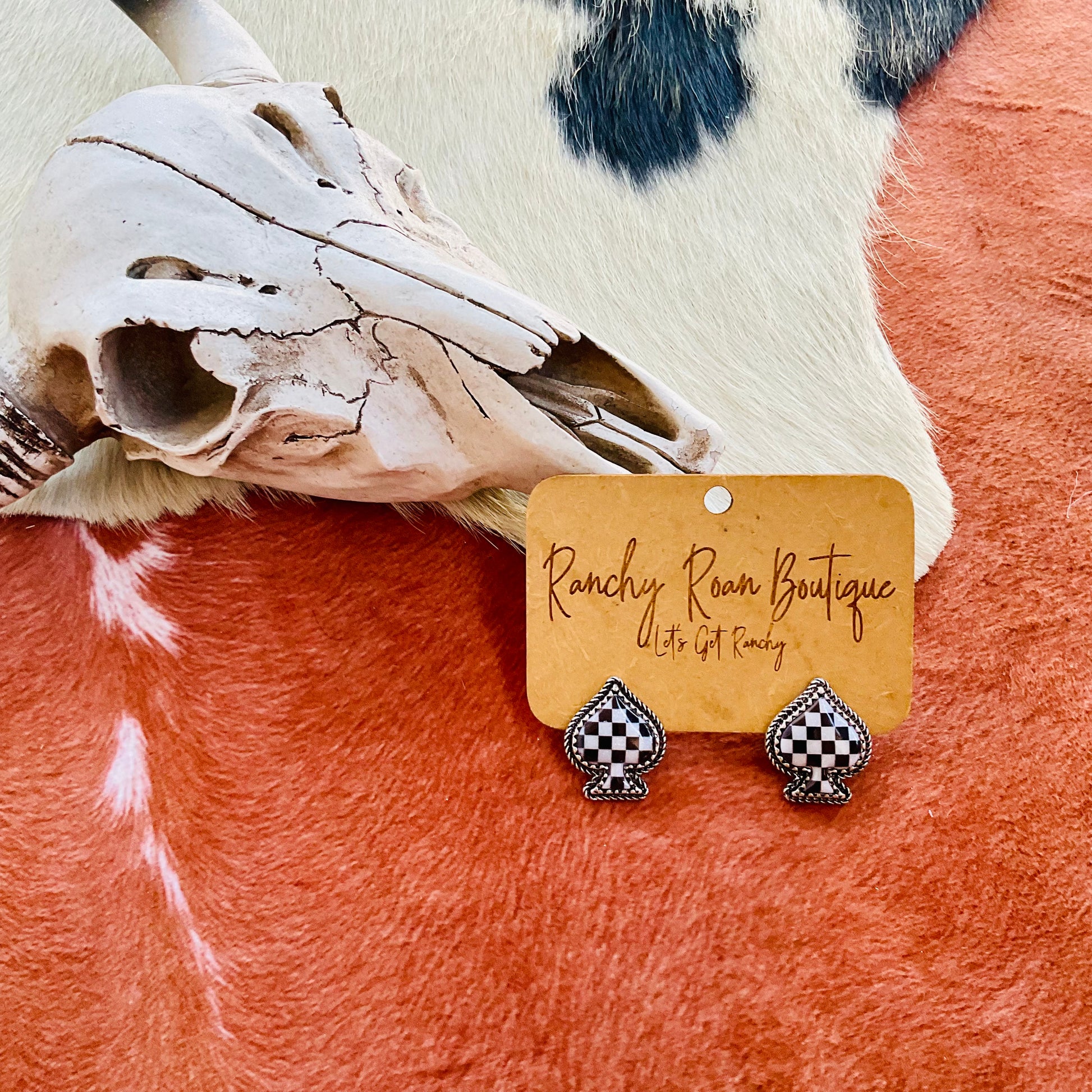 Checker-pattern spade-shaped stud earrings displayed on a Ranchy Roan Boutique tag, set against a Western-style cowhide background with a decorative skull.