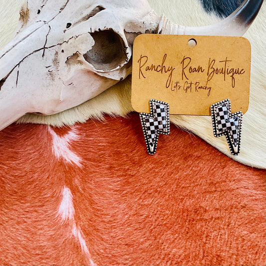 A pair of Checker Pattern Thunder Stud Earrings featuring a black and white checker pattern with burnished silver, displayed on a rustic cowhide background.