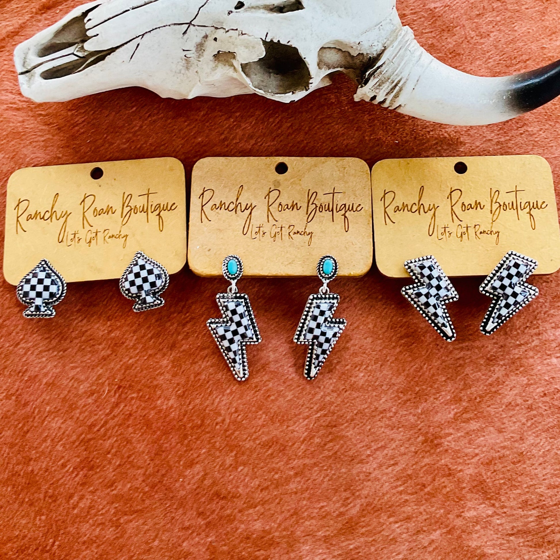 A set of checkered spade stud earrings and two pairs of checkered lightning bolt earrings displayed on Ranchy Roan Boutique cards next to a cow skull on a cowhide background.
