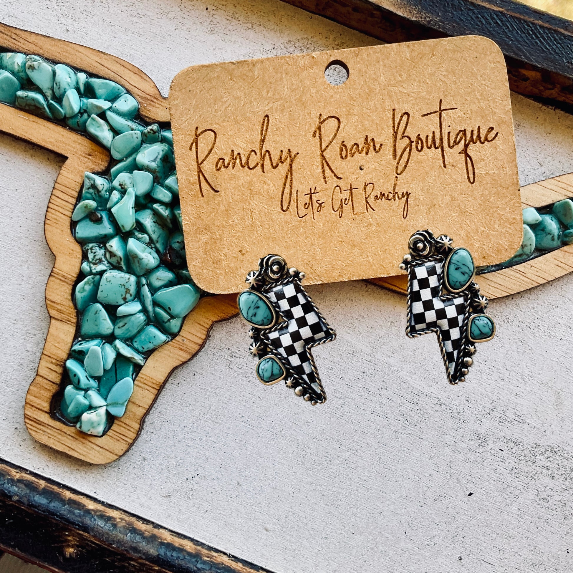 Checkered lightning bolt earrings with turquoise stones, displayed on a wooden turquoise-inlaid longhorn decor piece.