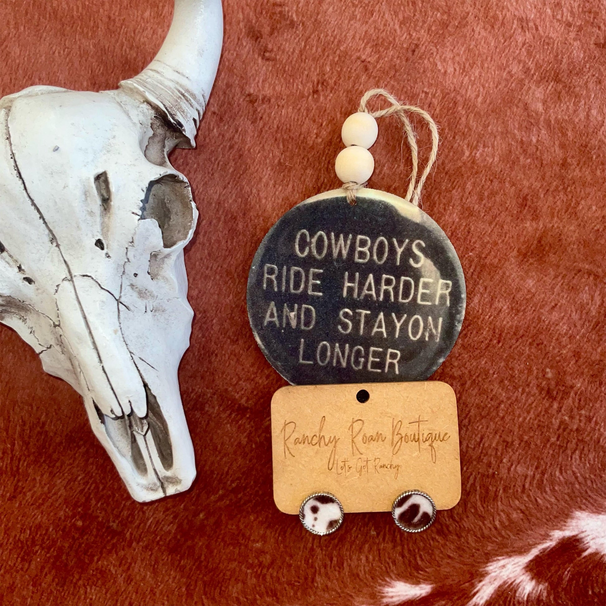 A round felt freshie with the message “Cowboys Ride Harder and Stay On Longer,” featuring a distressed design and wooden beads.