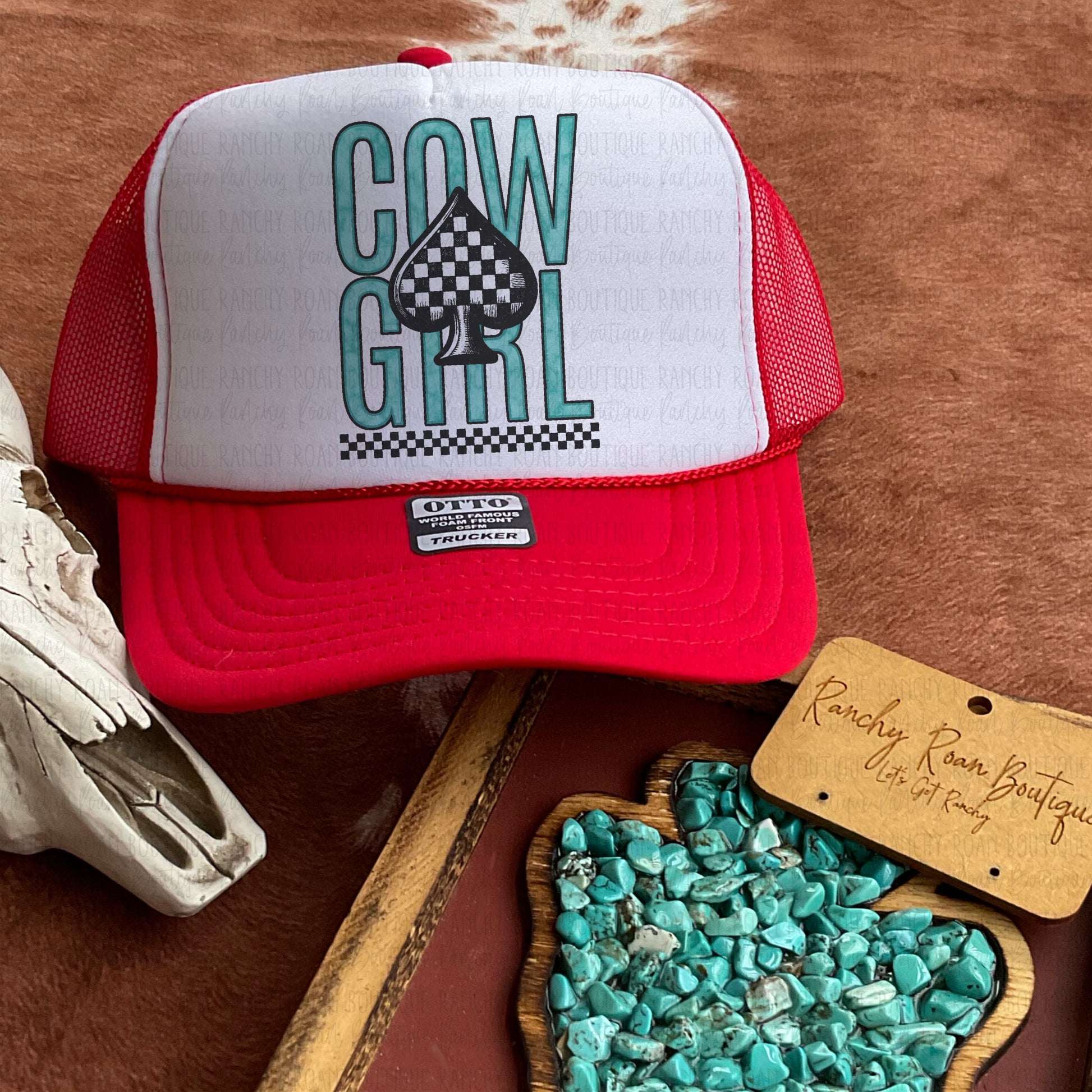 Red trucker hat with a white foam front featuring teal “Cowgirl” text and a black checkered spade graphic, styled with Western-themed decor.