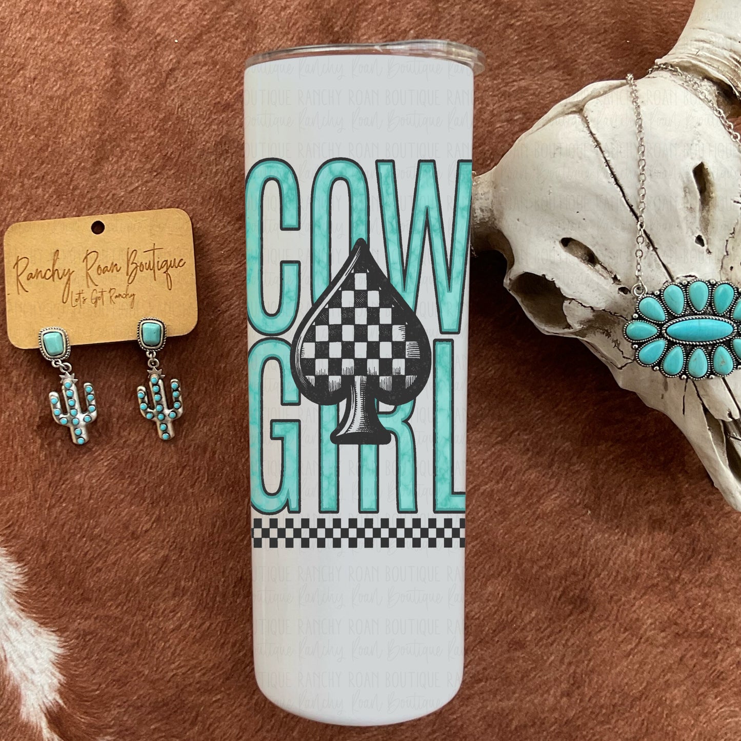 White tumbler featuring bold teal “Cowgirl” text with a checkered spade graphic, styled on a Western backdrop.