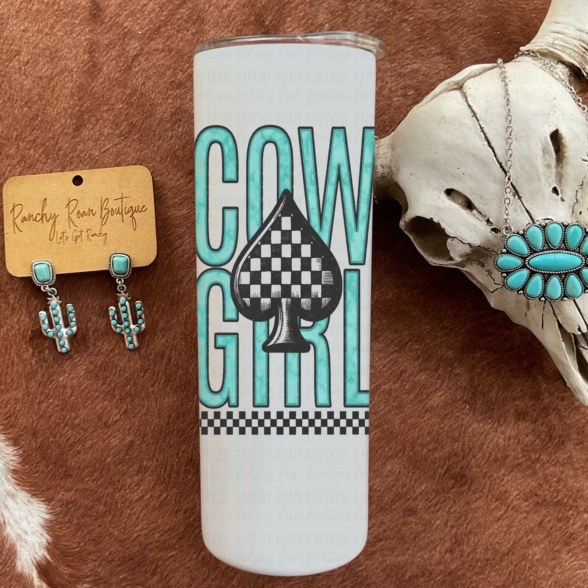 White tumbler featuring bold teal “Cowgirl” text with a checkered spade graphic, styled on a Western backdrop.