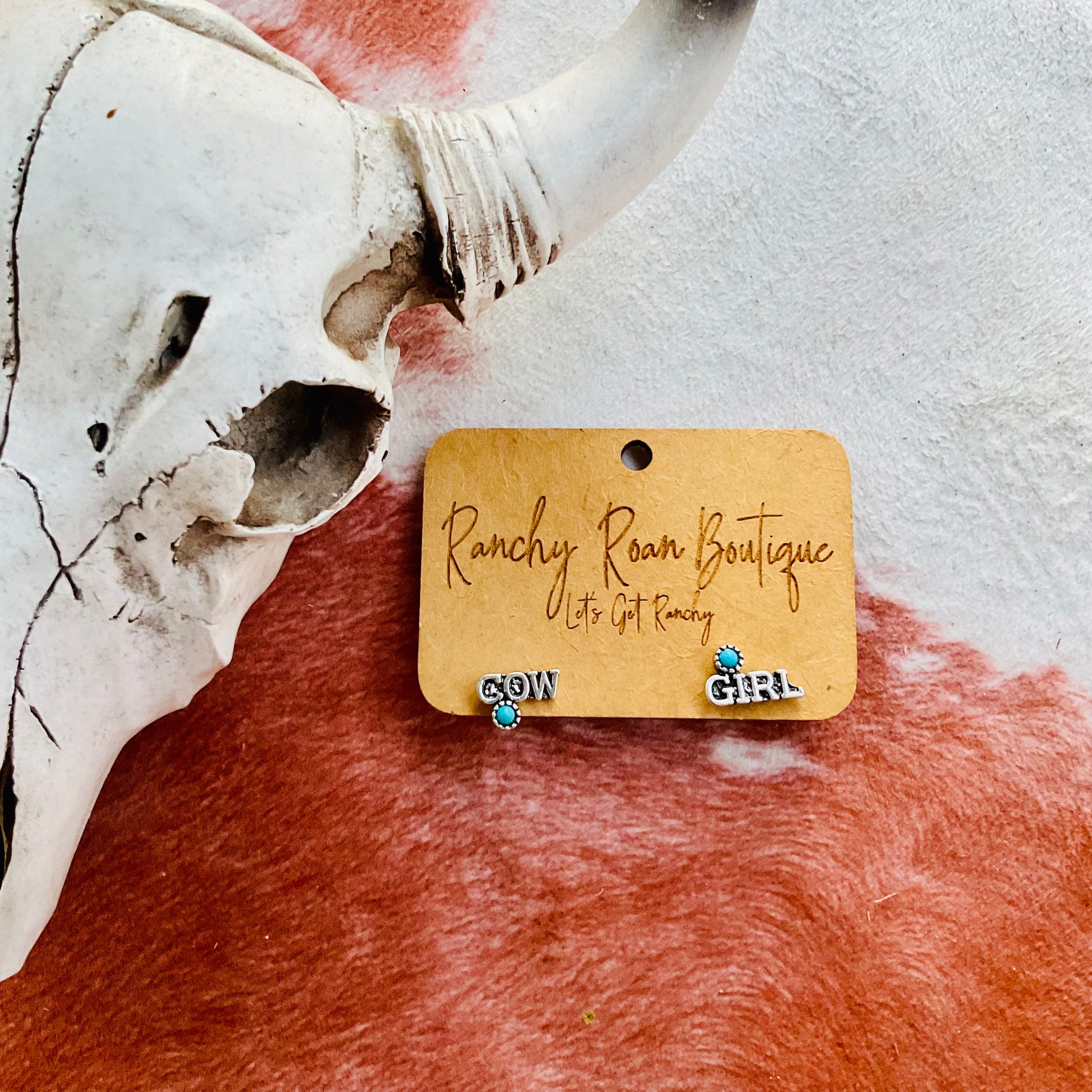 A pair of “COW” and “GIRL” word earrings with turquoise accents displayed on a Ranchy Roan Boutique card, set against a cowhide background with a decorative cow skull.