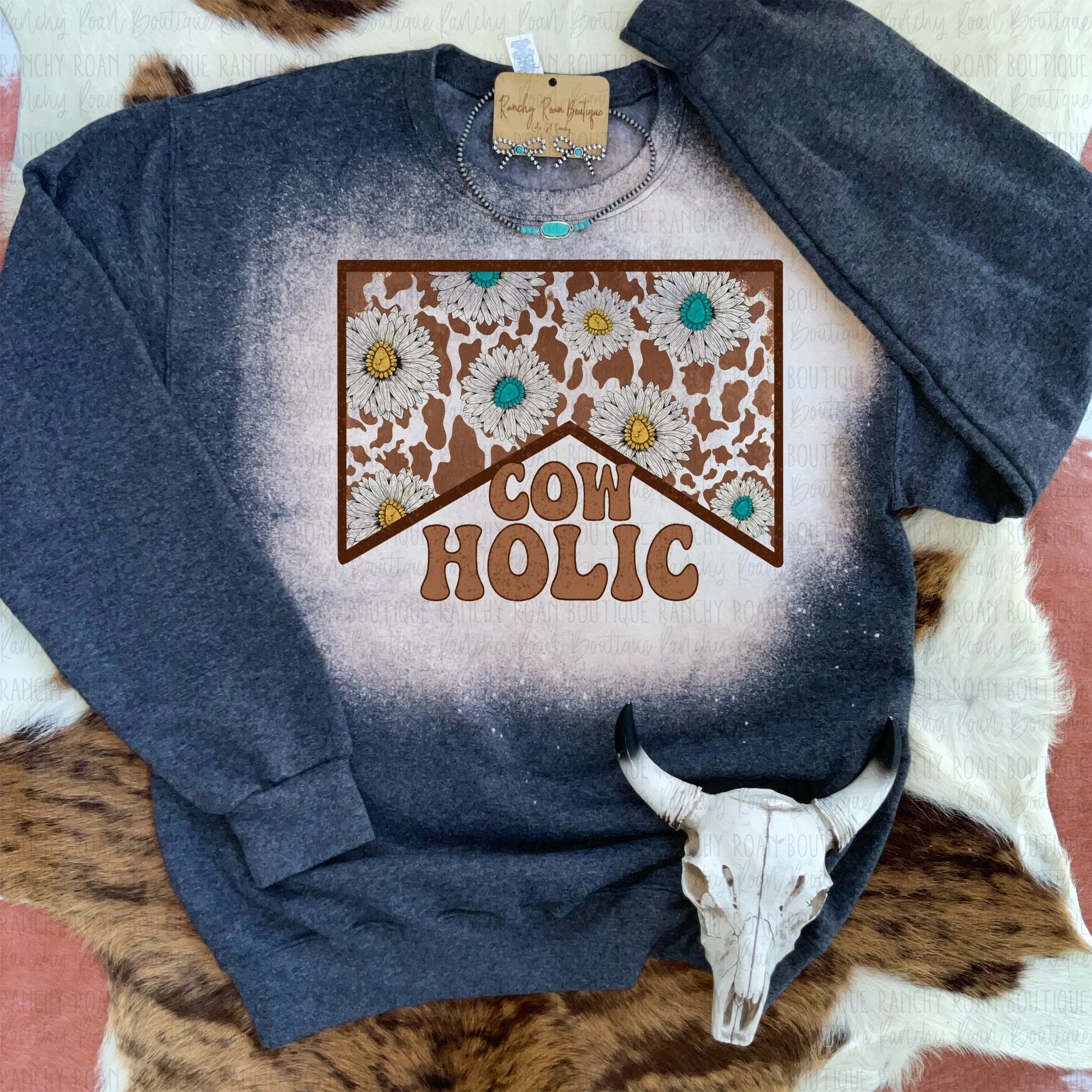 Dark heather sweatshirt with a “Cowholic” graphic, showcasing a cow print and floral accents, styled with Western-themed items.