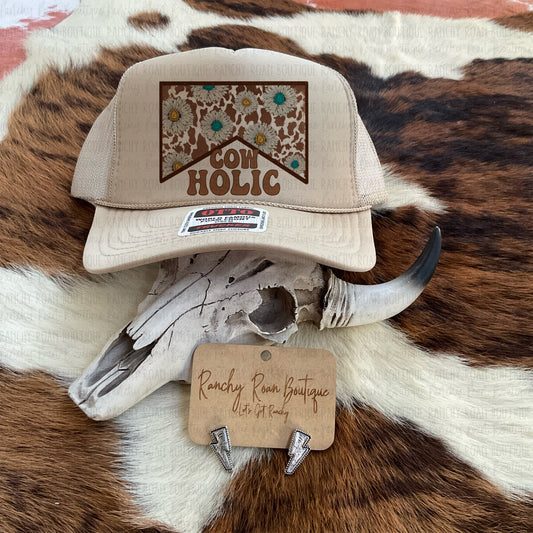 Tan trucker hat with “Cowholic” graphic featuring cow print and floral details, styled with Western accessories.