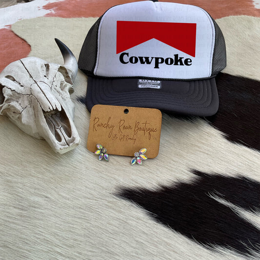 Black and white trucker hat with bold “Cowpoke” graphic in red and white, displayed with Western-themed decor.