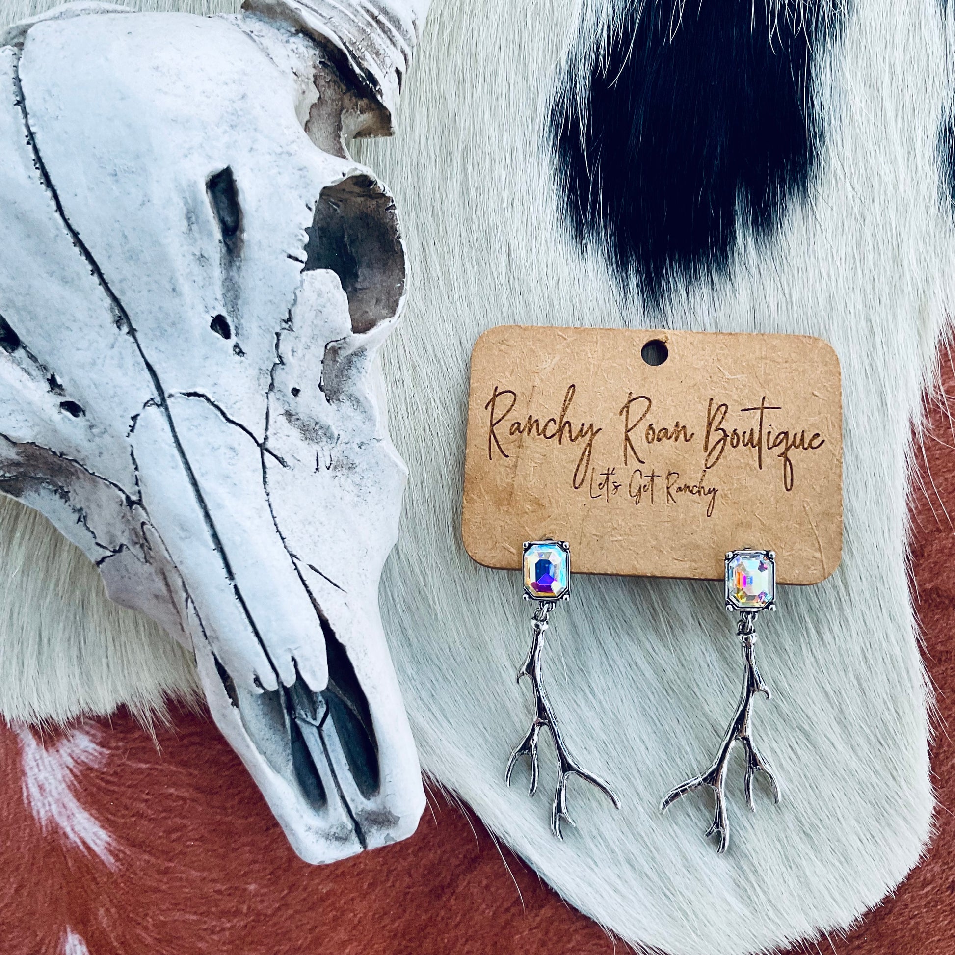 A pair of iridescent crystal stud earrings featuring silver antler dangles, displayed on Ranchy Roan Boutique branded cards against a cowhide background with a rustic cow skull for decoration.