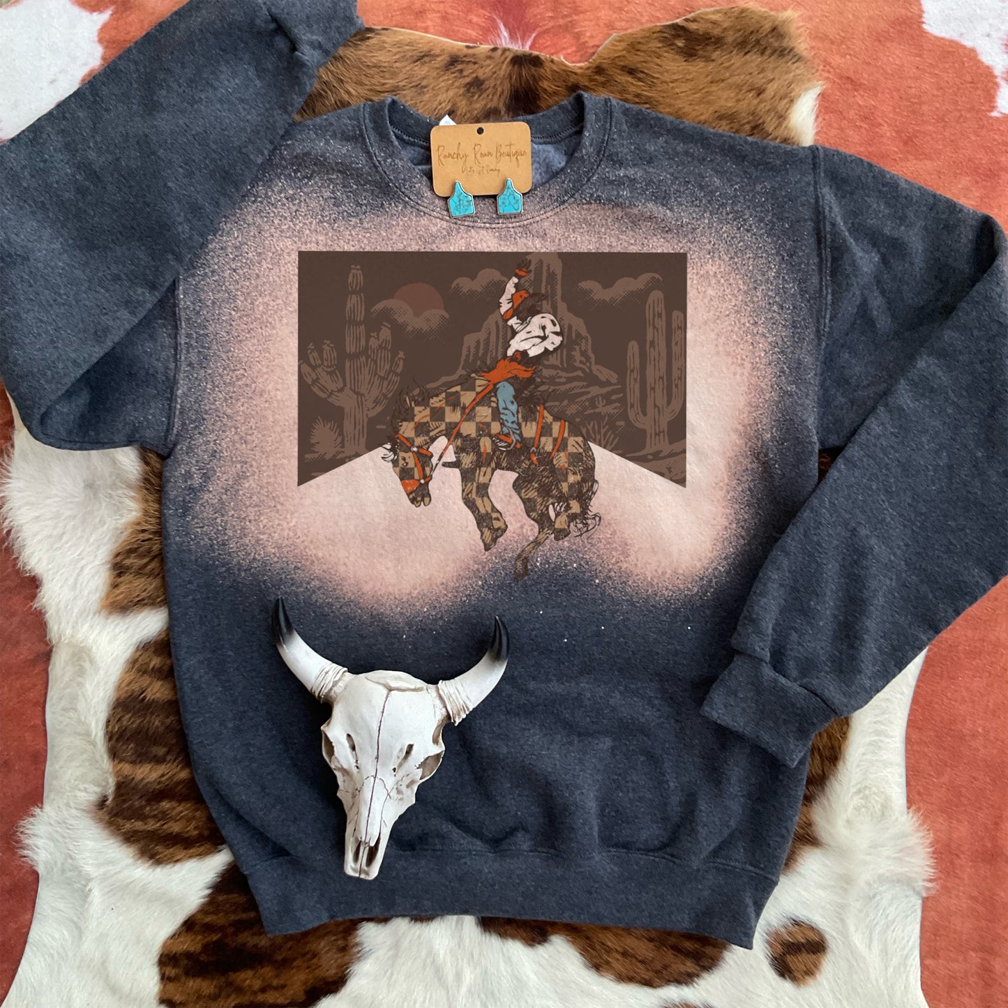 Desert Bucking Horse sweatshirt with bleached splatter detail displayed flat on a cowhide background, showcasing the Western design.