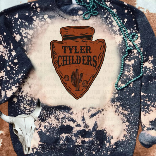 A bleached sweatshirt from Ranchy Roan Boutique featuring a large arrowhead design with the text “Tyler Childers” prominently displayed in the center. The sweatshirt has a unique bleached splatter pattern in shades of dark blue and cream, creating a one-of-a-kind look. The arrowhead graphic is rustic brown with cactus illustrations at the bottom, adding a Western touch. The sweatshirt is styled with a turquoise beaded necklace draped over it and a cow skull accessory placed nearby, enhancing the Western aes