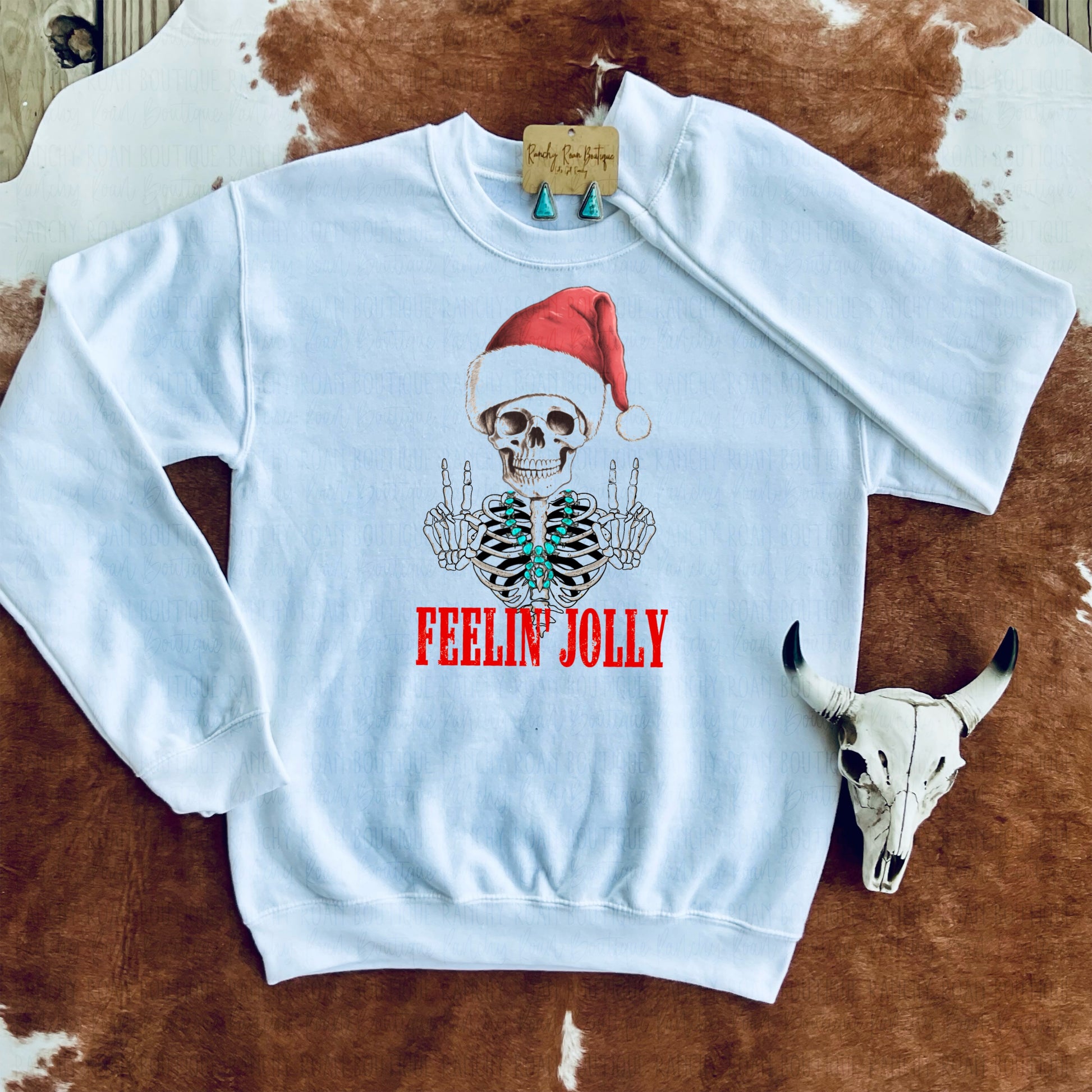 White sweatshirt featuring a skeleton wearing a Santa hat with the words “Feelin’ Jolly” in red, styled with a cow skull and turquoise earrings on a cowhide backdrop.
