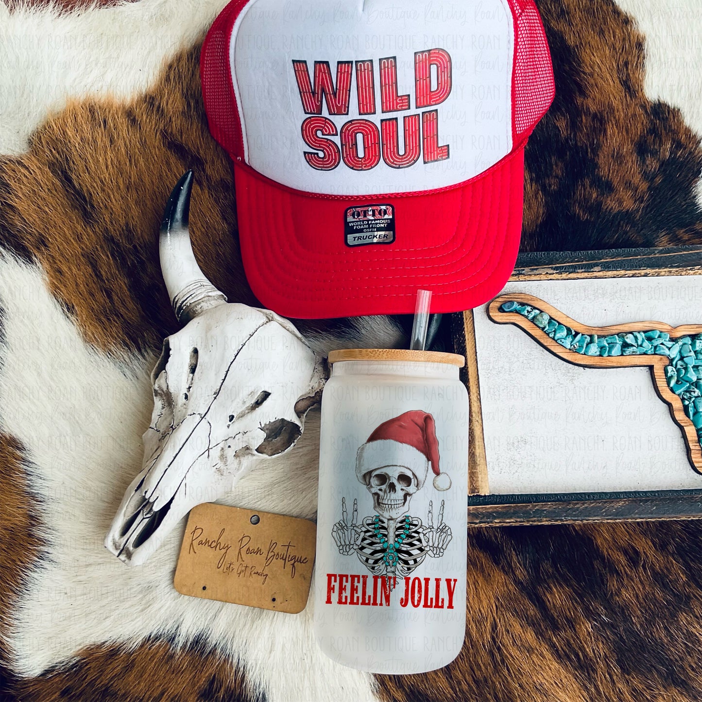 A glass tumbler featuring a skeleton wearing a Santa hat with the words “Feelin’ Jolly,” displayed with a cow skull and red trucker hat on a cowhide background