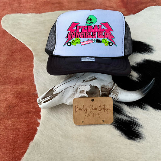 Trucker hat with neon skull and dice design, featuring “Feral Cowgirls Club” text, displayed on a cowhide with a decorative cow skull.