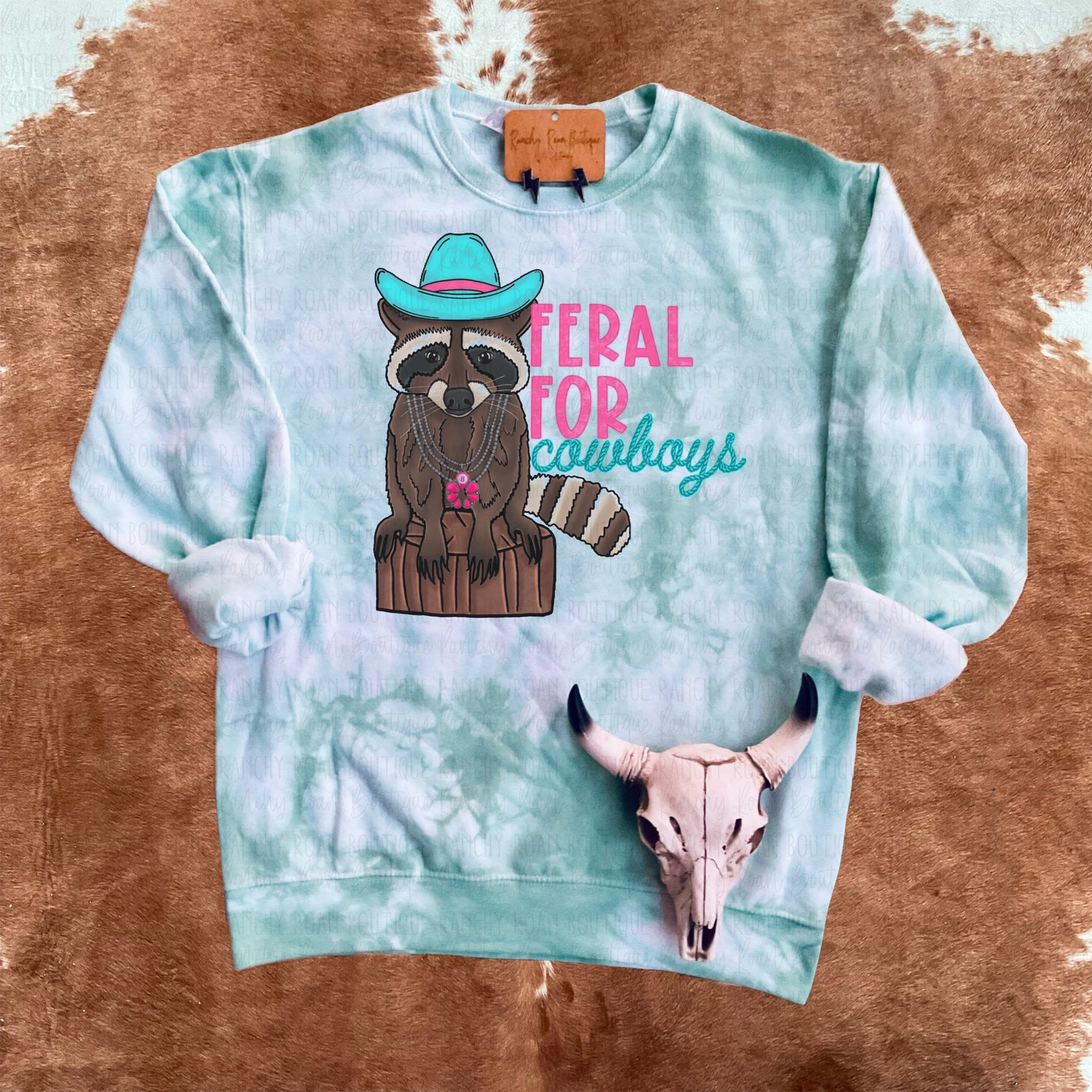 White sweatshirt dyed in varying shades of turquoise and green, featuring “Feral For Cowboys” sublimation graphic, displayed flat.