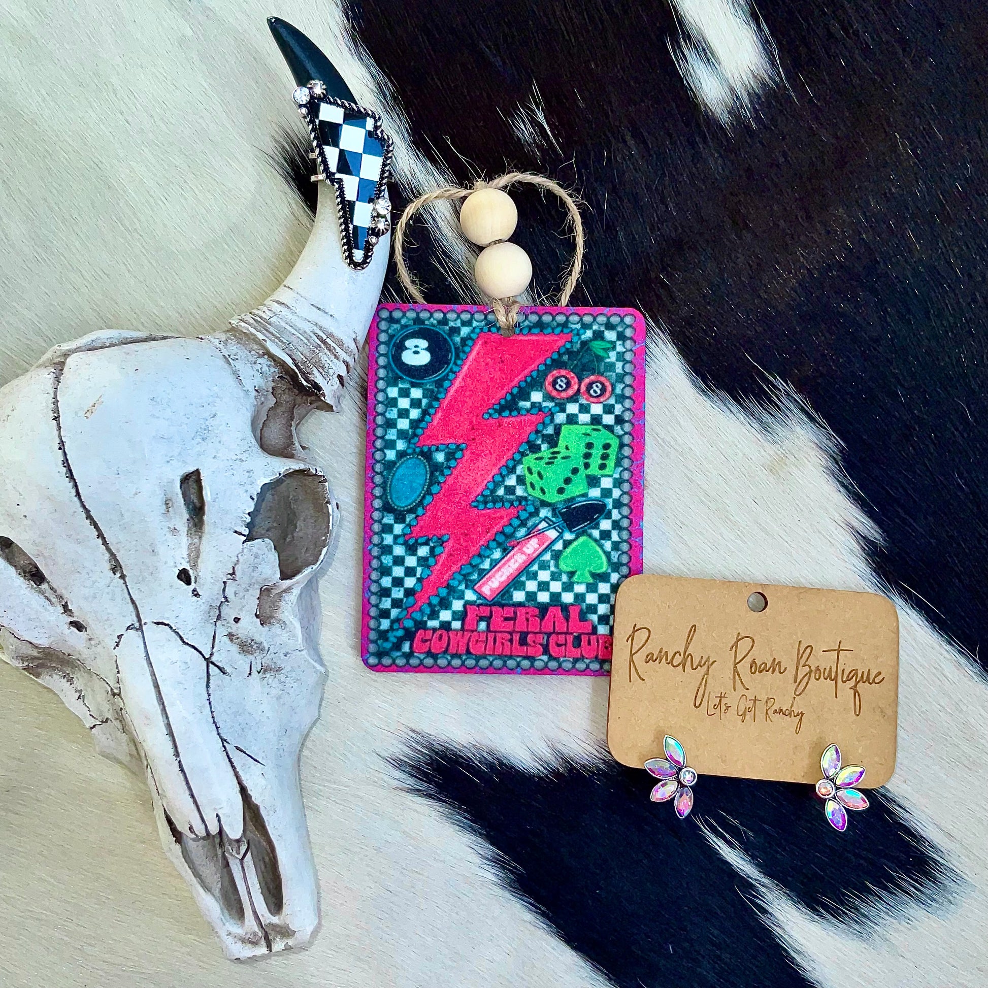 Feral Cowgirls Club felt air freshener featuring a neon lightning bolt, dice, and checkerboard pattern, styled on a cowhide background.