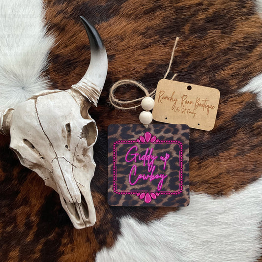 A square leather patch tag with pink script reading 'Giddy up Cowboy' and a pink border, displayed on a brown and white cowhide rug next to a skull and a wooden brand tag that reads 'Ranchy Rose Boutique'.