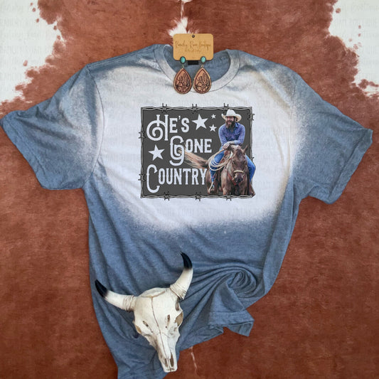 He’s Gone Country Western Graphic Tee featuring a cowboy on horseback with bold text and star accents, on a hand-bleached background, styled with rustic Western accessories.