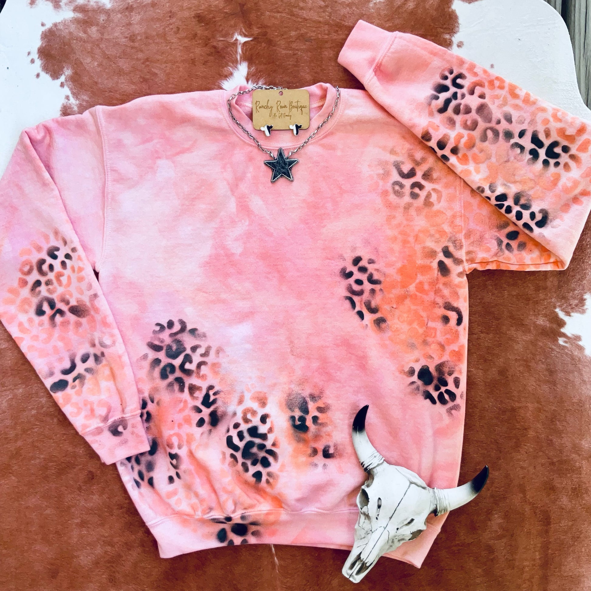 	Orange Leopard Distressed Western Sweatshirt with bold orange, pink, and red hues, featuring a distressed splatter leopard print design, styled with a star necklace on a rustic backdrop.