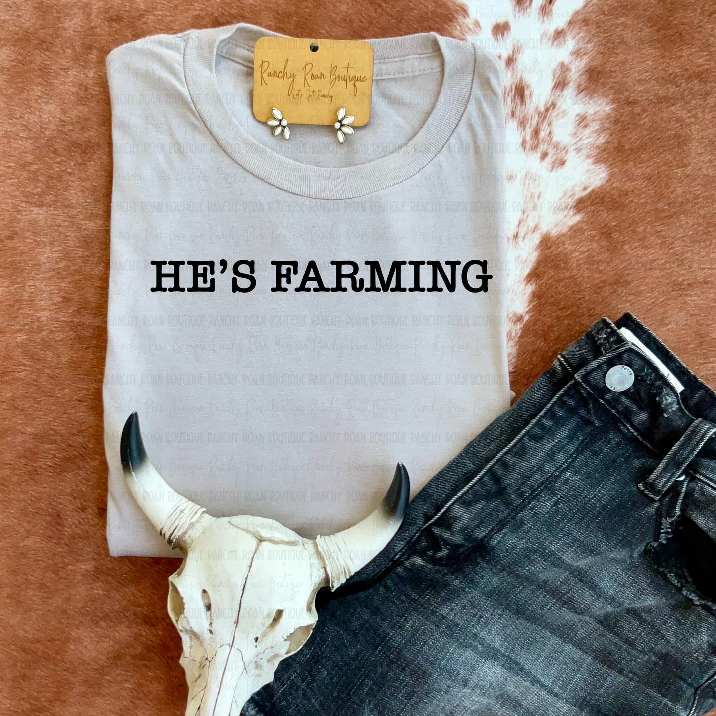Sand-colored sweatshirt with “He’s Farming” text folded flat on cowhide backdrop.